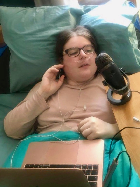 Kara Jane lies in bed, singing into a microphone on a table over her bed.