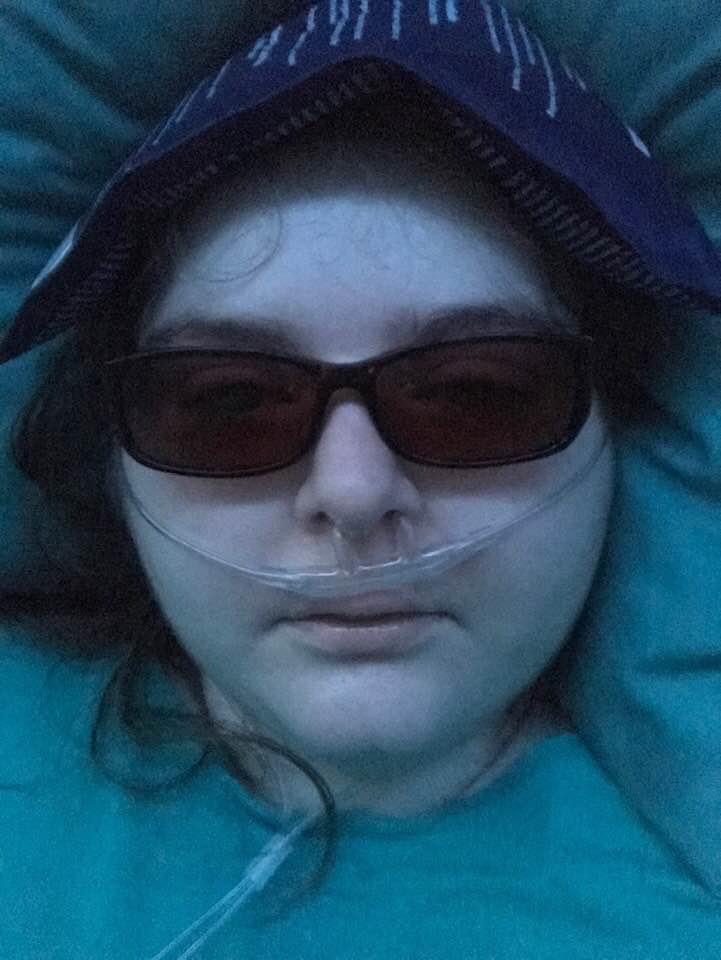 Kara wears dark glasses and has an ice pack on her head. She has an oxygen cannula in her nose. Her skin is pale and she looks very ill.