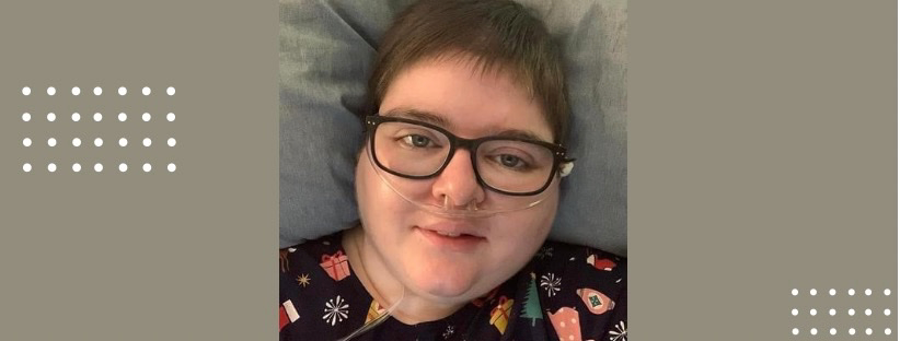 Close-up shot of Kara. She has cropped dark hair and wears glasses. She has an oxygen tube in her nose.